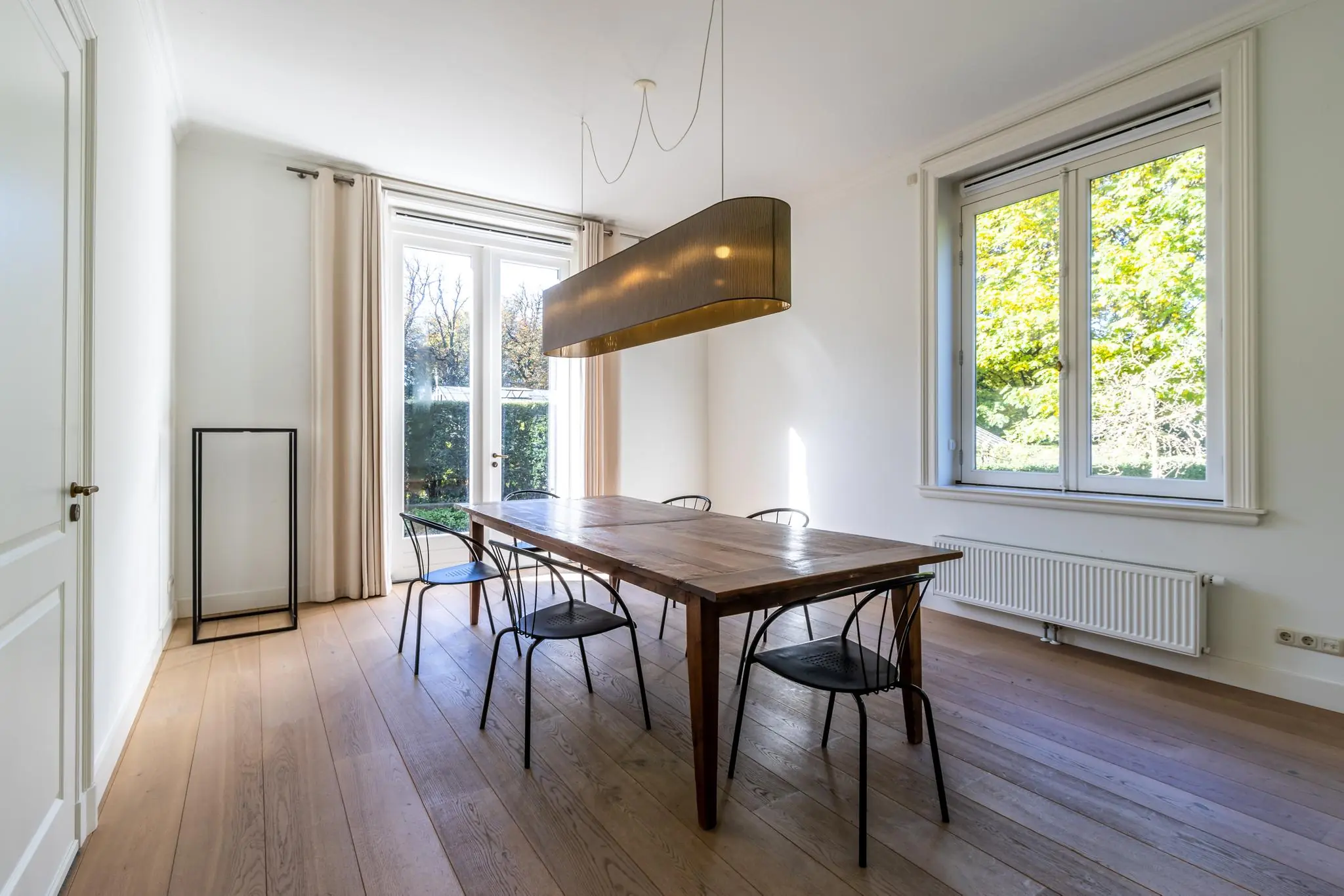 Lichte eetkamer aan de Langbroekerdijk A met houten vloer, minimalistische houten eettafel, zes zwarte stoelen en grote ramen met uitzicht op groen.